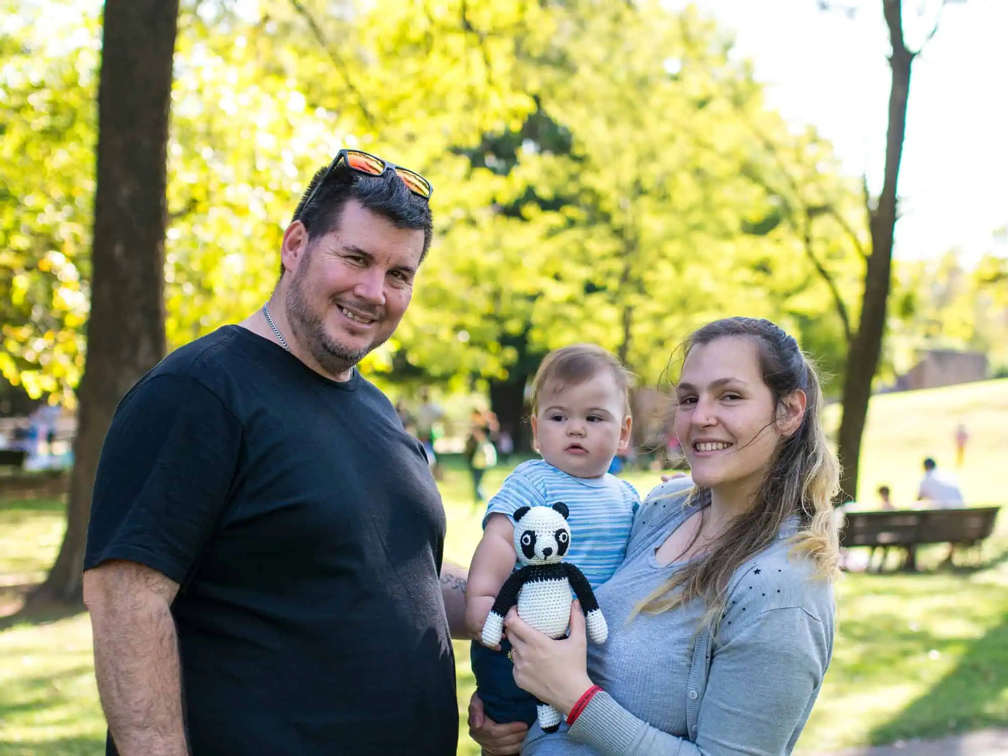 Glückliche Familie im Park: Mutter hält SindiBaba Panda-Kuscheltier, Baby auf dem Arm, Vater daneben.