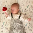 Babyjunge liegt auf gemusterter Decke und hält eine gehäkelte Fliegenpilz-Babyrassel