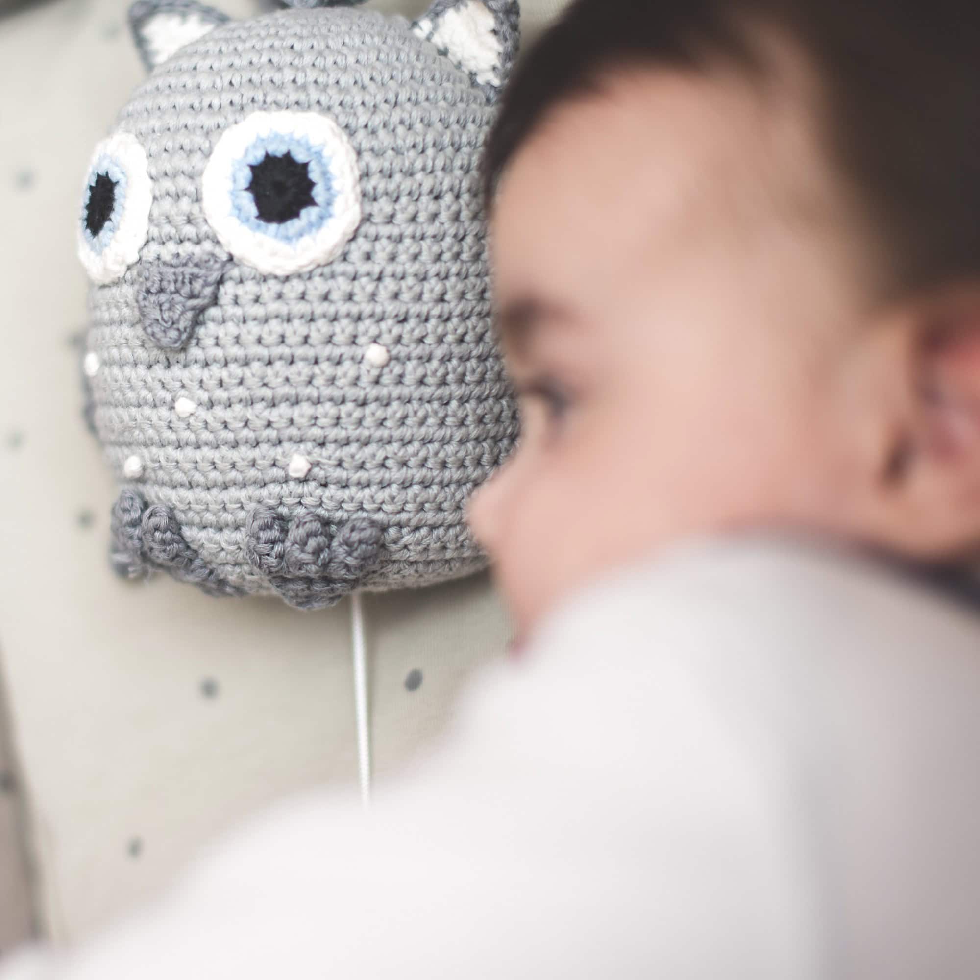 Müdes Kleinkind liegt im Bett neben der grauen SindiBaba Spieluhr „Eule“; Eule im Fokus, Kind weich unscharf im Vordergrund.